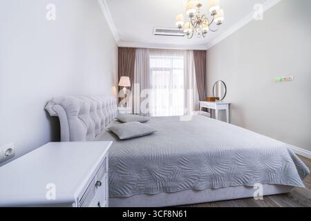 Una camera con una nuova e intima ristrutturazione in tonalità grigio-beige. Ampio letto matrimoniale con coperta grigia. Comodini e un tavolo in stile classico. Foto Stock