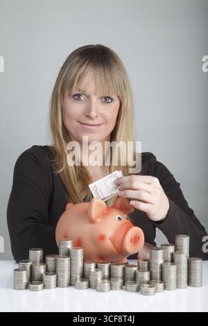 Una giovane donna che mette una banconota da dieci euro in un salvadanaio, davanti ha accumulato due monete in euro Foto Stock