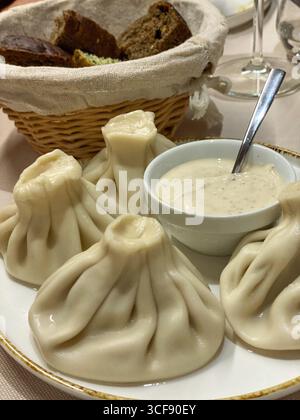 Gnocchi Khinkali appoggiati su un piatto di ceramica, conditi con panna acida alle erbe, cestino di pane appena sfornato nelle vicinanze, che mette in risalto la tradizione culinaria georgiana Foto Stock