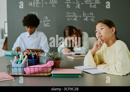 Ragazza asiatica che pensa mentre si siede con diversi adolescenti in classe Foto Stock