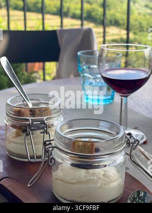 Due vasetti di dessert cremoso con biscotti e un bicchiere di vino rosso serviti su un tavolo con vista sulla campagna toscana Foto Stock
