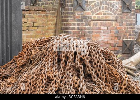 Un grande mucchio di catene arrugginite davanti a una classica parete in mattoni rossi con decorazioni in metallo, che mostrano texture e il passare del tempo in mezzo a un indaco Foto Stock