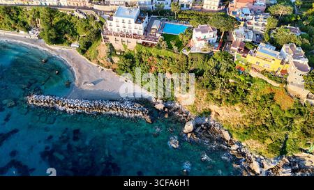 Case dai colori vivaci si aggrappano sulla collina e si affacciano su una tranquilla spiaggia dove le dolci onde si infrangono sulla riva. Un tramonto vibrante dipinge il cielo, Foto Stock
