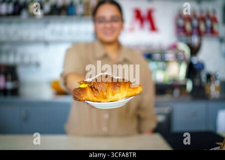 Cremona, Italia 25 luglio 2025 Barista serve un delizioso croissant appena sfornato ripieno di crema pasticcera e condito con zucchero granulato, offeri Foto Stock