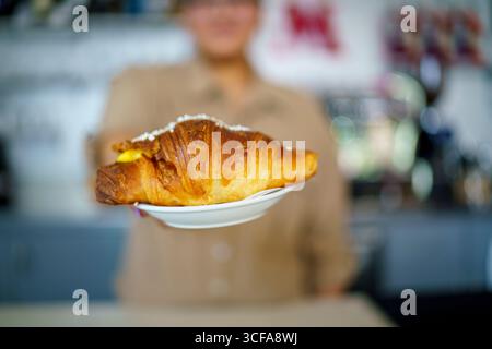 Cremona, Italia 25 luglio 2025 Barista serve un delizioso croissant appena sfornato ripieno di crema pasticcera e condito con zucchero granulato, offeri Foto Stock