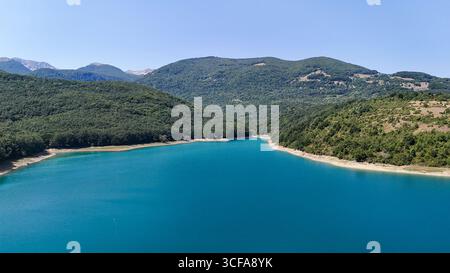 Sotto un radioso cielo di mezzogiorno, un tranquillo lago turchese riflette le maestose montagne circostanti. Alberi verdeggianti fiancheggiano la riva, creando un ambiente tranquillo Foto Stock