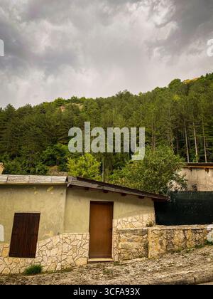 Dimora italiana con pareti in pietra e porte in legno intemperie, persiane rustiche che si affacciano sulla verdeggiante collina, annidate nel paesaggio di campagna di Rocca Pia Foto Stock