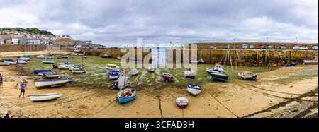 La bassa marea al porto di Mousehole in Cornovaglia, Regno Unito, rivela barche da pesca ormeggiate su fondali marini esposti, ideali per viaggi, pesca, stile di vita costiero, e. Foto Stock