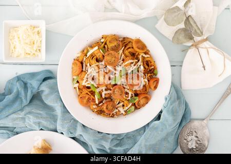 Pasta di pomodoro con erbe, pomodori ciliegini e formaggio - piatto vibrante e confortevole, preparato su piatto bianco con panno blu e cucchiaio. Foto Stock