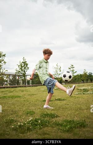 Ragazzo caucasico che calcia la palla da calcio su un campo erboso, concentrandosi sul bilanciamento della palla con un piede, ambiente all'aperto con alberi e cielo nuvoloso sullo sfondo Foto Stock