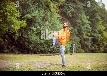 Uomo barbuto in giacca a vento che si allunga ginocchio-petto in parco con palo per esercizi, spazio per fotocopie Foto Stock