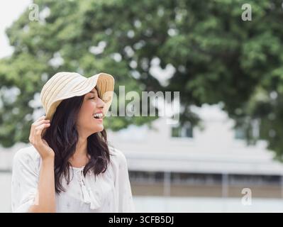 Donna ispanica sorridente mentre indossa una camicetta bianca che tocca il cappello di paglia all'aperto, copia spazio Foto Stock