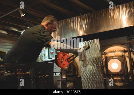 Vetro fuso rosso brillante sul tubo di soffiaggio sopra il banco dell'officina in mezzo a forni, utensili, condotti di ventilazione Foto Stock