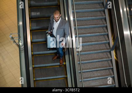 Uomo in piedi sulla scala mobile con guida in vetro al terminale che tiene la valigia nel blazer, spazio fotocopie Foto Stock