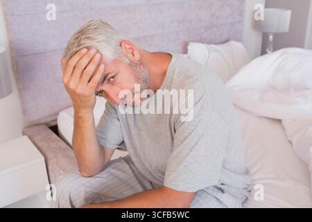 Uomo di mezza età seduto sul bordo del letto premendo la fronte accanto alla lampada del comodino con pigiama grigio Foto Stock