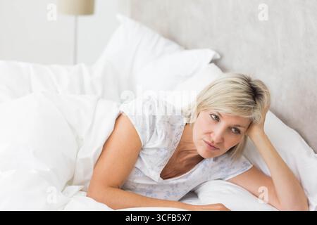 Donna di mezza età sdraiata sul letto in camera da letto, appoggiata sul piumone bianco sotto la lampada del comodino Foto Stock