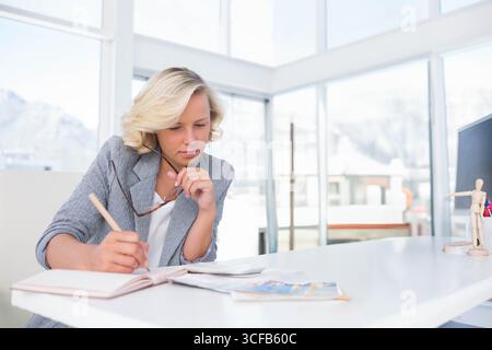 Donna professionista che indossa un blazer grigio che scrive su un notebook e tiene gli occhiali alla scrivania dell'ufficio Foto Stock