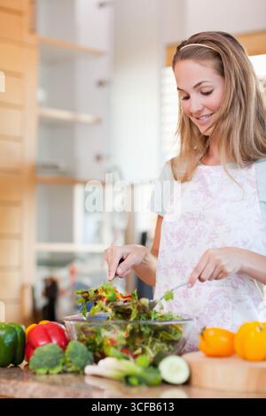 Donna che indossa un grembiule floreale che getta verdi a foglia in una ciotola di vetro trasparente sul piano di lavoro della cucina Foto Stock