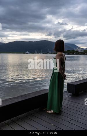 retro di donna che guarda al mare con un abito verde che posa la passerella di legno sul mare, una donna elegante che indossa un abito verde contro una scenografica bac sul mare Foto Stock