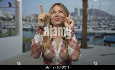Donna bionda con un sorriso allegro punta entrambe le dita indice verso l'alto al molo del porticciolo; ottimismo di anticipazione. Foto Stock