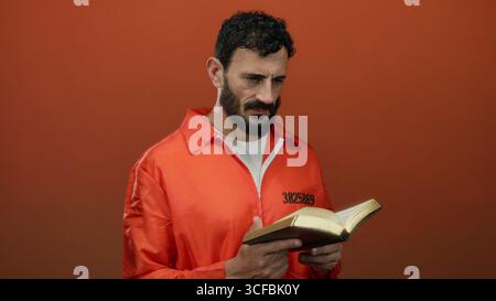 L'uomo ispanico con barba in tuta da prigione arancione tiene la bibbia su sfondo arancione, simboleggiando la riflessione e la redenzione. Foto Stock