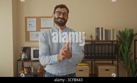 L'uomo ispanico che indossa occhiali e barba applaude entusiasticamente in uno spazio ufficio con un microonde e documenti su una tavola da stiro. Foto Stock