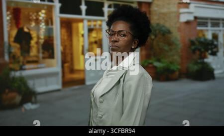 Donna sicura di sé con gli occhiali si staglia su una strada della città di fronte a una boutique di notte, mostrando un ambiente elegante e urbano all'aperto. Foto Stock