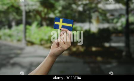 Mano che regge la bandiera svedese in un ambiente cittadino all'aperto che simboleggia l'orgoglio nazionale e la cultura. Foto Stock