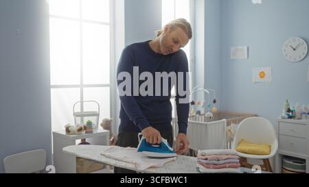 L'uomo maturo che stirava nella lavanderia interna di casa si concentra sulle faccende domestiche circondate da giocattoli per bambini che mettono in risalto la vita domestica e la responsabilità Foto Stock
