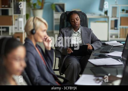 Giovane donna adulta di colore che indossa le cuffie sedute alla scrivania tenendo gli appunti che lavora in un ufficio moderno con due colleghi in primo piano utilizzando computer e cuffie Foto Stock