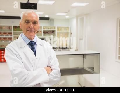 Scienziato senior che indossa un camice da laboratorio in piedi con le braccia incrociate in laboratorio accanto ai vasetti per campioni Foto Stock