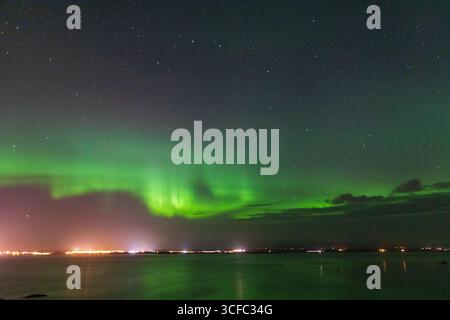 Vista delle affascinanti sfumature verdi e bianche dell'aurora boreale danzano attraverso il cielo notturno sopra le calme acque di Vik, Islanda. Foto Stock