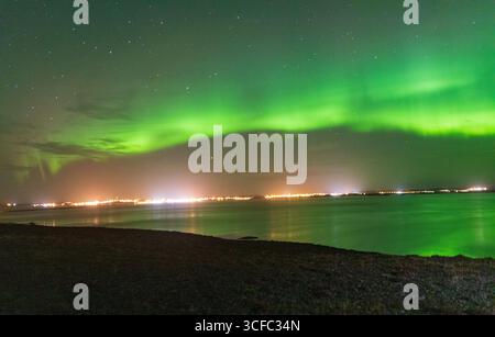 Vista delle scintillanti danze verdi dell'aurora boreale sopra le lontane luci della città, che si specchiano nelle acque tranquille e scure, un balletto celeste di luce e natura, Vik, Islanda. Foto Stock