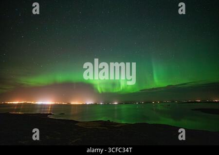 Vista delle luminose danze verdi dell'aurora boreale sopra le acque scure che riflettono le lontane luci della città, creando uno spettacolo etereo, Vik, Islanda. Foto Stock