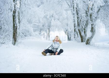 Donna seduta su un sentiero innevato circondato da alberi innevati. Indossa una giacca d'argento, sorridendo pacificamente, godendosi il tranquillo paesaggio invernale. A. Foto Stock