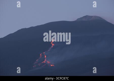 Nell'agosto del 2025 l'Etna entrò in una fase effusiva alimentata da bocche tra bocca nuova e il cratere sudorientale a 2.980–3.200 m s.l.m., inviando freschi corsi di lava lungo la base del cratere sudorientale e sui fianchi sudovest/occidentali superiori. Le immagini mostrano turisti curiosi e gruppi guidati che osservano i flussi luminosi e le esplosioni stromboliane intermittenti al cratere sudorientale, con piccole scogliere di cenere e pennacchi di gas che si disperdono rapidamente. Altri sfiati annotati il 20 e il 21 agosto, con flussi che avanzano da sud a sud-ovest attraverso l'area della cima. Foto Stock