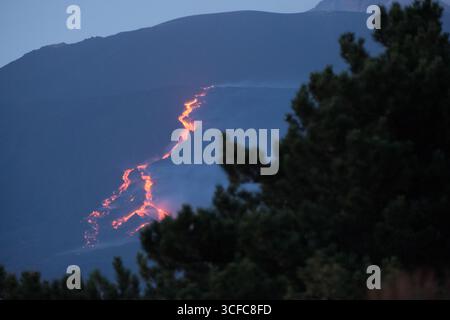Nell'agosto del 2025 l'Etna entrò in una fase effusiva alimentata da bocche tra bocca nuova e il cratere sudorientale a 2.980–3.200 m s.l.m., inviando freschi corsi di lava lungo la base del cratere sudorientale e sui fianchi sudovest/occidentali superiori. Le immagini mostrano turisti curiosi e gruppi guidati che osservano i flussi luminosi e le esplosioni stromboliane intermittenti al cratere sudorientale, con piccole scogliere di cenere e pennacchi di gas che si disperdono rapidamente. Altri sfiati annotati il 20 e il 21 agosto, con flussi che avanzano da sud a sud-ovest attraverso l'area della cima. Foto Stock