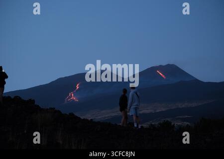 Nell'agosto del 2025 l'Etna entrò in una fase effusiva alimentata da bocche tra bocca nuova e il cratere sudorientale a 2.980–3.200 m s.l.m., inviando freschi corsi di lava lungo la base del cratere sudorientale e sui fianchi sudovest/occidentali superiori. Le immagini mostrano turisti curiosi e gruppi guidati che osservano i flussi luminosi e le esplosioni stromboliane intermittenti al cratere sudorientale, con piccole scogliere di cenere e pennacchi di gas che si disperdono rapidamente. Altri sfiati annotati il 20 e il 21 agosto, con flussi che avanzano da sud a sud-ovest attraverso l'area della cima. Foto Stock
