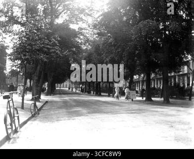 Gloucestershire, Inghilterra. Circa 1905 - una fotografia edoardiana della Promenade, Cheltenham. I pedoni camminano lungo i marciapiedi e i ciclisti stanno utilizzando la strada. Un uomo sta camminando lungo la strada indossando un panino che pubblicizza una vendita di ombrelloni a Kendalls. Questa azienda, situata a Cheltenham High Street, è specializzata in ombrelloni, bastoni da passeggio e ombrelloni. Alla fine della passeggiata si trova il Queen's Hotel. Foto Stock