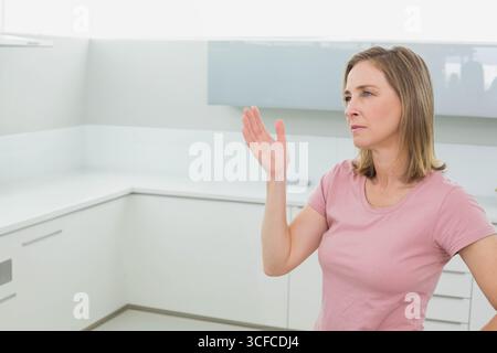Donna adulta matura che solleva la mano destra in una cucina minimalista con armadi bianchi e spazio per le copie Foto Stock