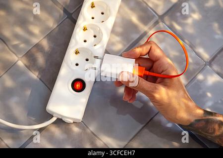 Donna che collega il caricabatterie a filo elettrico alla presa multipla, sicurezza energetica domestica per la ricarica dei dispositivi elettrici. Foto Stock