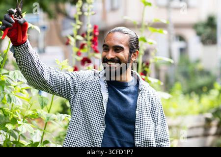 Uomo che giareggia all'aperto, sorridendo mentre cura le piante in un lussureggiante giardino urbano. Germania Foto Stock