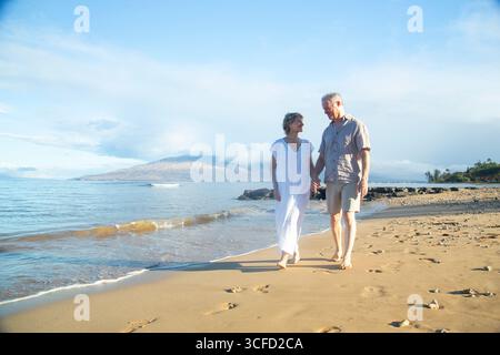 Una coppia di anziani cammina mano nella mano su una spiaggia soleggiata, lasciando impronte nella sabbia dorata. Parco Kepaniwai, Wailuku, Hawaii Foto Stock