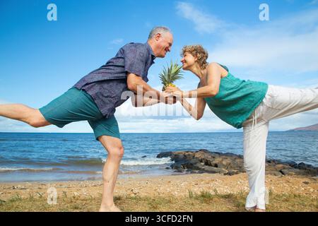 Coppia senior in equilibrio su una gamba vicino alla spiaggia, condividendo un momento di gioco con un ananas. Parco Kepaniwai, Wailuku, Hawaii Foto Stock