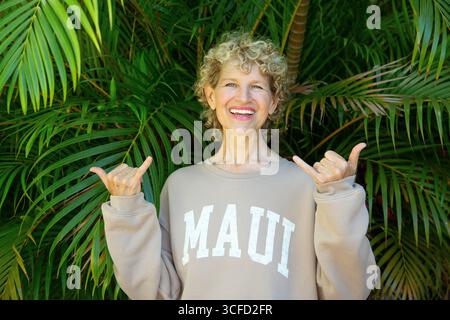 Donna sorridente in felpa beige con testo Maui, in piedi davanti a foglie verdi lussureggianti. Parco Kepaniwai, Wailuku, Hawaii Foto Stock