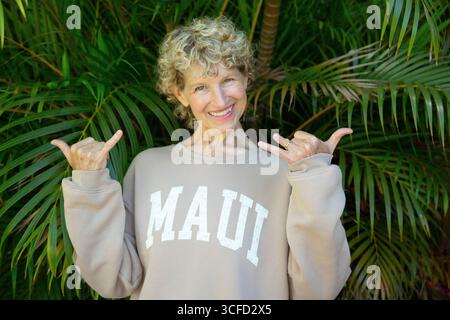 Donna sorridente con capelli ricci in una felpa Maui che gestisce un cartello shaka davanti alle foglie di palma. Parco Kepaniwai, Wailuku, Hawaii Foto Stock