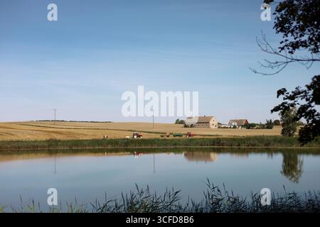 Vista panoramica di un lago rurale polacco. Campi falciati, attrezzature agricole e agricole durante la formazione del fieno visibili in lontananza Foto Stock