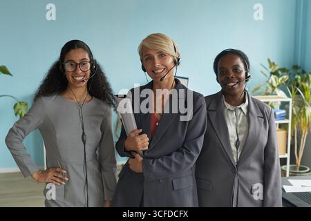 Tre diverse donne che indossano le cuffie, sorridono davanti alla fotocamera, dimostrando il lavoro di squadra in un moderno ambiente d'ufficio Foto Stock