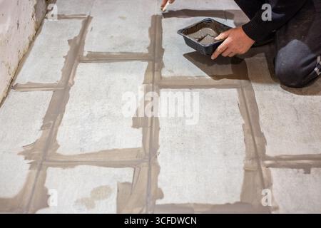 Un lavoratore applica la Malta tra piastrelle di pavimento di grandi dimensioni appena posate utilizzando un contenitore piccolo per il materiale. Foto Stock