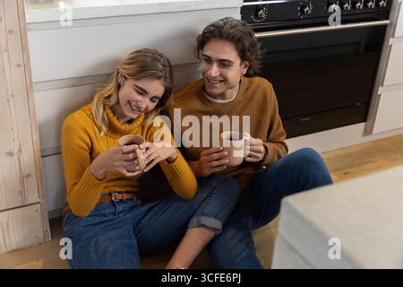 Indossa maglioni in maglia seduti sul pavimento della cucina appoggiati contro gli armadi e tenendo tazze in ceramica Foto Stock